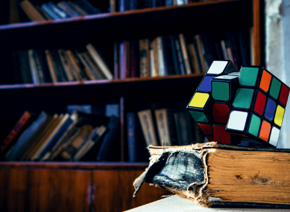 A rubiks cube balances on top of an old book, with a shelf of books in the background.