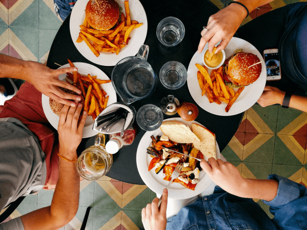 healthy eating image 3 A top down view of four friends eating out at a cafe. Their food is on the table: 3 burgers with chips and one person eating roasted vegetables with bread.